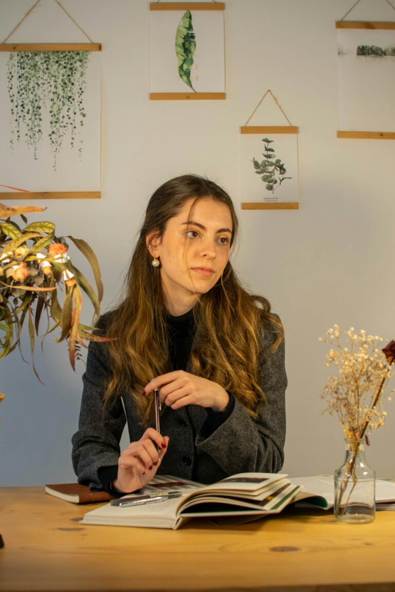A young woman pensively writing at her desk in a cozy home office filled with plants and art.