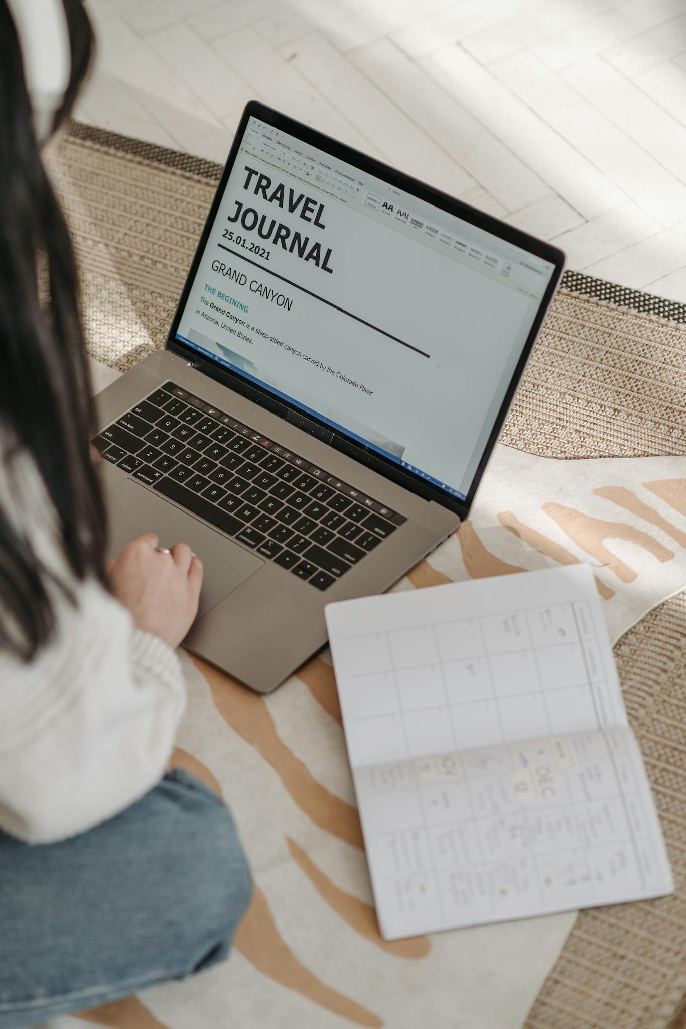 A young woman plans a travel journal using a laptop and notebook indoors on a cozy rug.