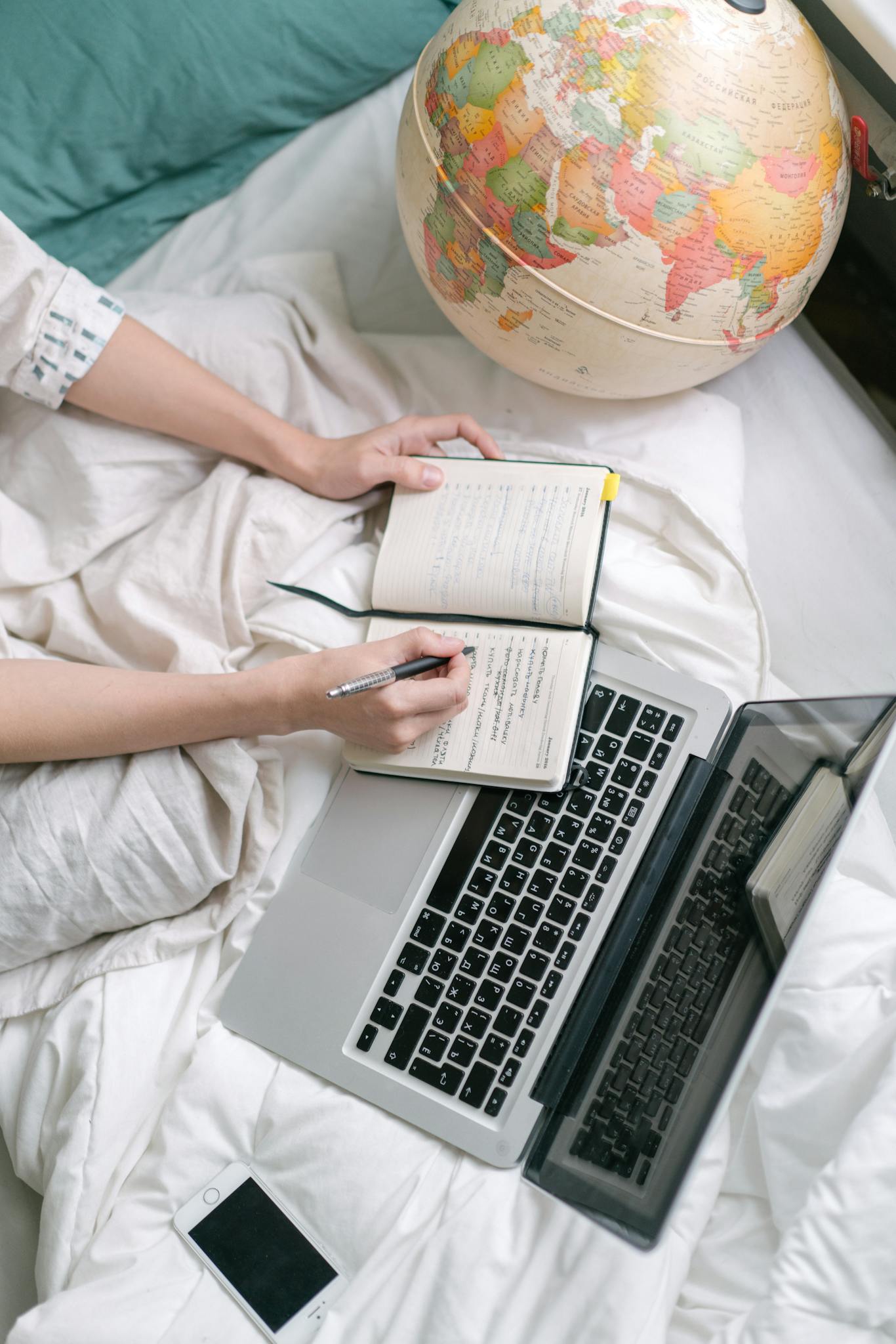 Overhead view of a cozy workspace with a laptop, notebook, and globe on a bed.